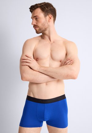 Homme musclé portant un boxer bleu, debout les bras croisés, regardant vers sa gauche sur un fond clair uni.