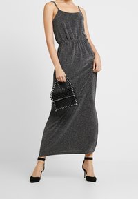 Robe maxi noire scintillante avec des bretelles spaghetti et une taille élastique. Associée à un sac géométrique noir orné de clous et des chaussures à talons hauts.