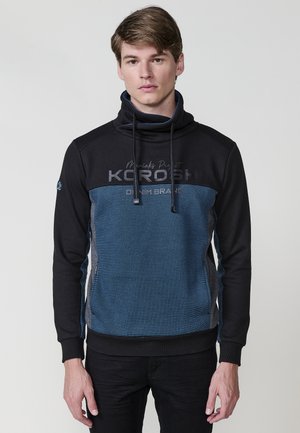 Blauer und schwarzer Pullover-Hoodie mit hohem Kragen, ausgestattet mit einem strukturierten Muster, Logo auf der Brust und kontrastierenden grauen Seitenpaneelen.
