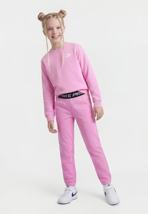 Niña sonriente con cabello rubio trenzado en dos moños, vistiendo sudadera rosa de Nike, pantalones deportivos a juego y zapatillas blancas de Nike, de pie sobre fondo blanco.