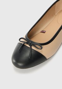 Ballerine beige avec un embout arrondi noir et un détail nœud noir sur le dessus sur un fond clair.
