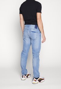 Jeans de denim azul claro con corte regular, con bolsillos traseros y una etiqueta azul oscura en la cintura, combinados con zapatillas blancas y negras.