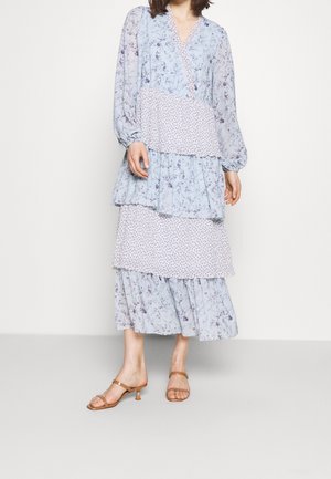 Robe fleurie bleue à design superposé, avec manches longues et motifs variés. Associée à des talons à brides marron clair.