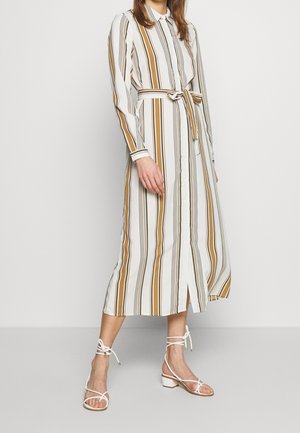 Kobieta w białej, zapinanej na guziki długiej sukience z pionowymi musztardowymi i czarnymi paskami, z przewiązanym paskiem oraz białe sandały na obcasie z wiązaniem wokół kostki.