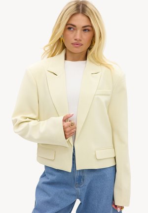 Femme blonde portant un blazer crème oversize, un top blanc et un jean bleu, avec des boucles d'oreilles créoles dorées et des bagues, regardant légèrement sur le côté.