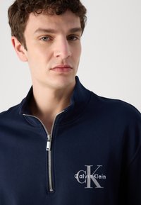 Felpa con zip blu navy e colletto alto, caratterizzata da una texture a coste e un logo Calvin Klein bianco sul petto. Dettaglio della zip in argento.