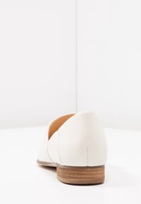 Chaussure plate en cuir blanc avec un bout arrondi et un petit talon en bois empilé. Texture lisse avec des détails de couture subtile à l'arrière.