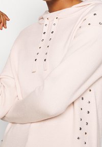 Mjukt rosa hoodie med metalliska nitar längs snoddarna, axlarna och sidorna, bärs av en person med armarna i kors.