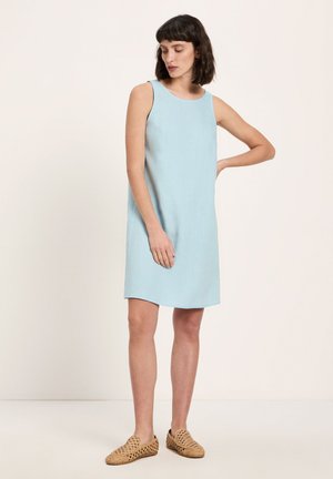 Woman with short dark hair wearing a sleeveless light blue knee-length dress and beige woven slip-on shoes, standing with one hand on hip.