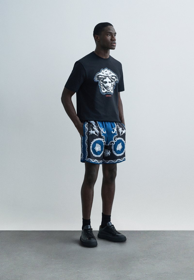 Black T-shirt with a graphic head design, paired with blue patterned shorts. Shoes are black, with a smooth texture. Minimalist setting.