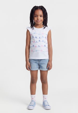 Νεαρή κοπέλα με πλεξούδες, φορώντας λευκό αμάνικο τοπ με την επιγραφή "Guess 1981 USA", τζιν σορτς, λευκές κάλτσες με ροζ καρδιές και ανοιχτό μπλε αθλητικά παπούτσια.