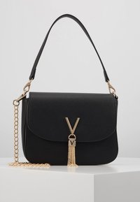 Svart syntetisk handväska med klaffstängning, guldkedja som axelrem och en dekorativ tofs med en metallisk V-emblem. Slät textur, rektangulär form.