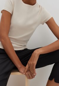 Beige ribbad kortärmad topp med rund halsringning, texturerad tyg och figurnära design, matchad med svarta byxor. Händerna vilar på en träpall.