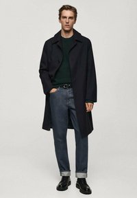 Črn trench coat preko zelene puloverke, v kombinaciji z zvijanimi temno modrimi jeans hlačami in črnimi čevlji. Enostaven dizajn z gumbi.