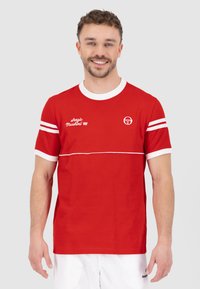 Rode katoenen T-shirt met witte accenten en strepen. Voorzien van geborduurd "Sergio Tacchini '66" logo op de voorkant. Korte mouwen en een ronde hals.