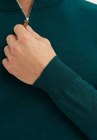 Pull en teal avec zip et poignets côtelés, présentant une languette en cuir sur la fermeture éclair. Le tissu semble lisse et léger.
