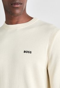 Sudadera de cuello redondo color beige claro, hecha de una tela suave, con un logo negro de "BOSS" en el pecho y detalles acanalados en las mangas.