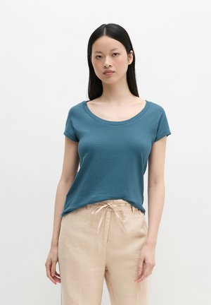 Kobieta w turkusowym krótkim rękawie z dekoltem w serek i beżowych spodniach ze sznurkiem, stojąca na jednolitym białym tle.