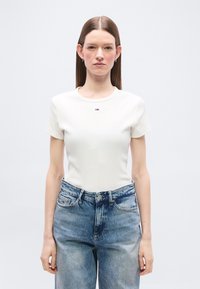 Γυναίκα με ίσια καστανά μαλλιά φοράει λευκό εφαρμοστό τοπ με κοντά μανίκια και ψηλόμεσα, άνετα μπλε τζιν, στέκεται μπροστά σε ένα απλό φωτεινό φόντο.