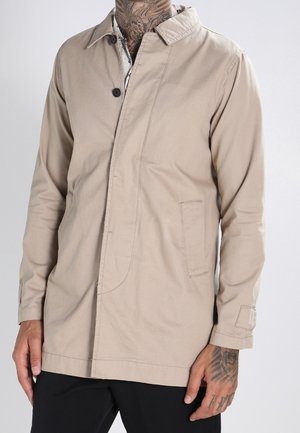 Veste beige boutonnée portée sur une chemise à col, avec des tatouages visibles sur le cou et les mains.