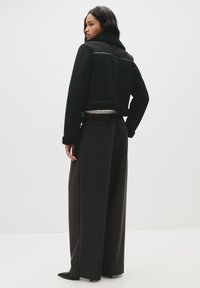 Giacca nera corta con colletto in shearling e dettagli in pelle, abbinata a pantaloni scuri a vita alta e gamba larga, e scarpe nere a punta.