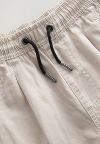 Helle beigefarbene Shorts aus einem strukturierten Stoff, mit elastischem Bund und schwarzen Kordeln mit silbernen Spitzen. Sichtbare Nähdetails.