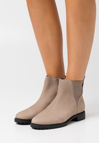 Botas de ante topo a la altura del tobillo con suelas de goma negras, usadas con las piernas desnudas sobre un fondo blanco.