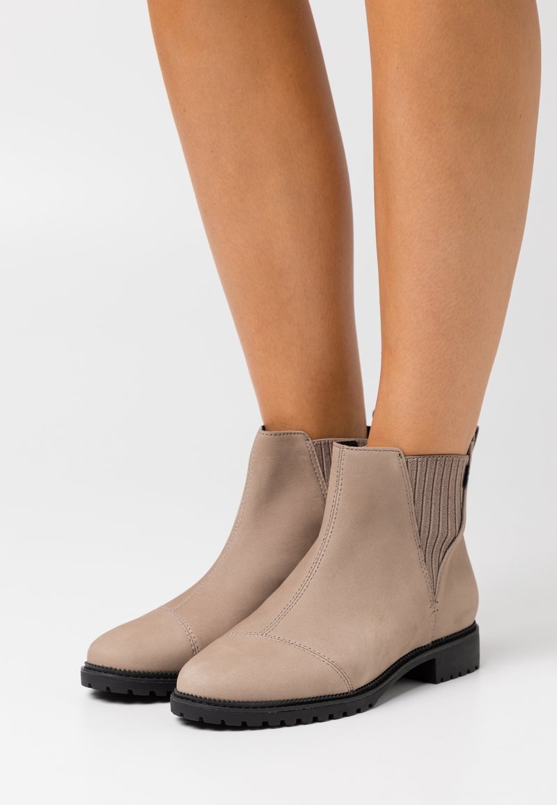Botas de ante topo a la altura del tobillo con suelas de goma negras, usadas con las piernas desnudas sobre un fondo blanco.