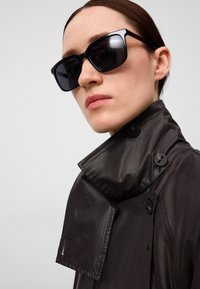 Donkerbruine, lichte trenchcoat met een hoge kraag en knoopaccenten. Model draagt oversized zwarte zonnebrillen, wat de geometrische vormen benadrukt.