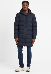 Man standing wearing a navy padded hooded coat, dark blue jeans, and white sneakers against a plain white background.