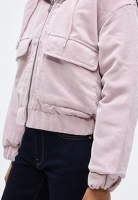 Ροζ κοντό ζακέτο από κορδόνι με δύο τσέπες στο στήθος, ριμπ υφή, ελαστικές μανσέτες κ αι κεντρικό κλείσιμο με φερμουάρ. Ορατό μπλε τζιν παντελόνι.