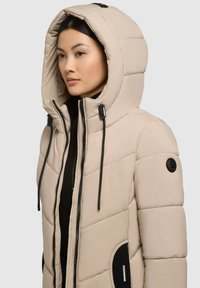 Svetlobéžová prešívaná bunda s vysokým golierom a nastaviteľnou kapucňou so sťahovacími šnúrkami, šikmým prešívaním a čiernym logom.