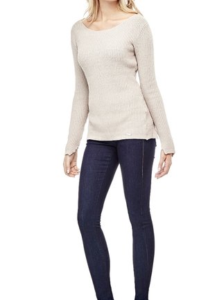 Femme portant un pull beige côtelé à manches longues et un jean skinny bleu foncé, debout avec les bras détendus le long du corps.