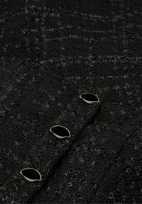 Zwarte stof met textuur en een houndstoothpatroon, met in het midden vier zwarte ovale knopen met metalen accenten.