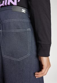 Pantalones de mezclilla oscura con costuras blancas, que presentan un bolsillo trasero y una etiqueta de cuero negro con un logotipo. Mano descansando sobre la tela.