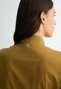 Chemise marron à col, avec une texture lisse, mettant en valeur un petit accent doré au dos du col. Les détails comprennent un design minimaliste.