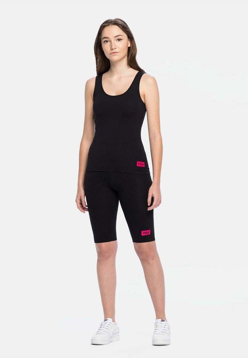 Schwarzes Tanktop und Radshorts aus dehnbarem Material mit einem pinkfarbenen Logo-Patch an der Seite. Das Model steht vor einem schlichten Hintergrund.