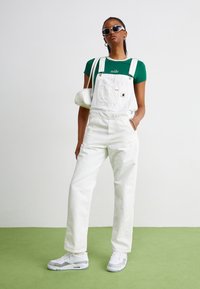 Salopette in denim bianco con spalline regolabili, abbinata a una maglietta verde con logo bianco. Indossata con sneakers bianche e una piccola borsa bianca.