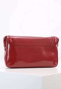 Borsa a tracolla rettangolare rossa lucida con chiusura a pattina, appoggiata su una superficie bianca contro uno sfondo bianco uniforme.