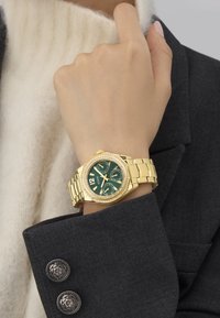 Ceas de aur cu un cadran verde și accente de pietre strass, având o brățară groasă și un carcasă octogonală, purtat pe o mână aproape de un pulover și un sacou.