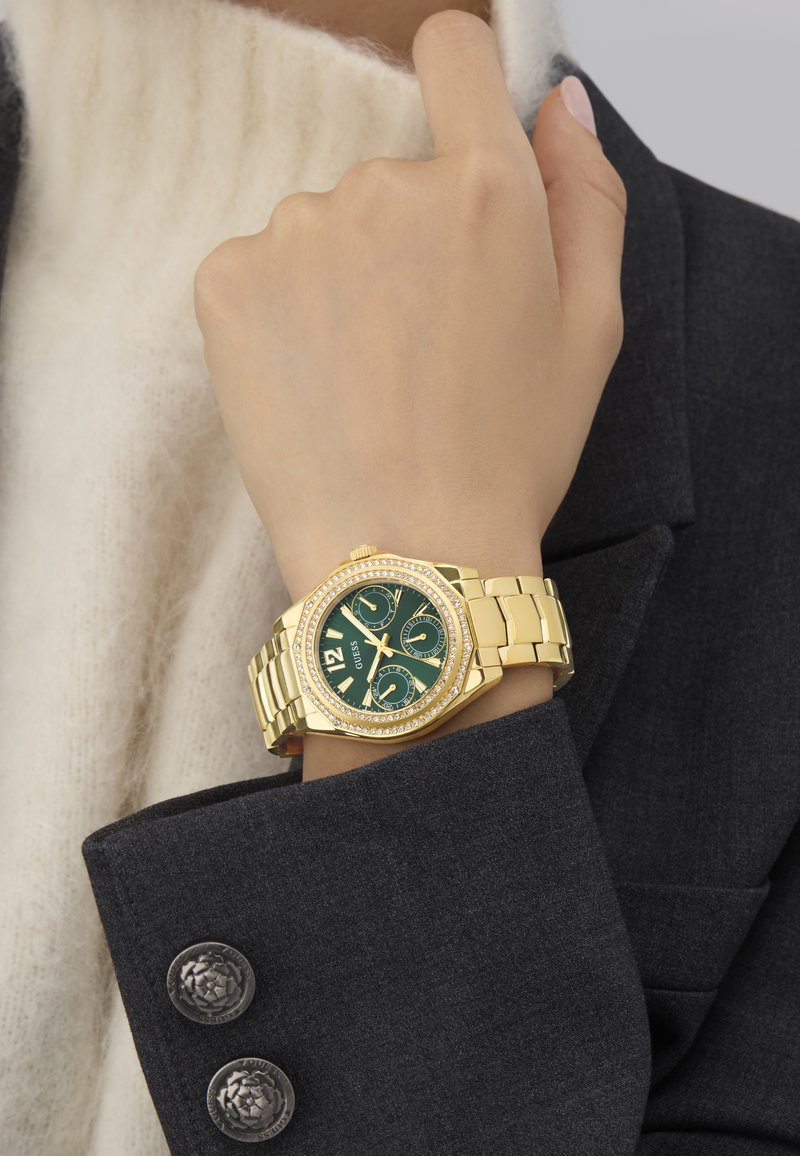 Ceas de aur cu un cadran verde și accente de pietre strass, având o brățară groasă și un carcasă octogonală, purtat pe o mână aproape de un pulover și un sacou.