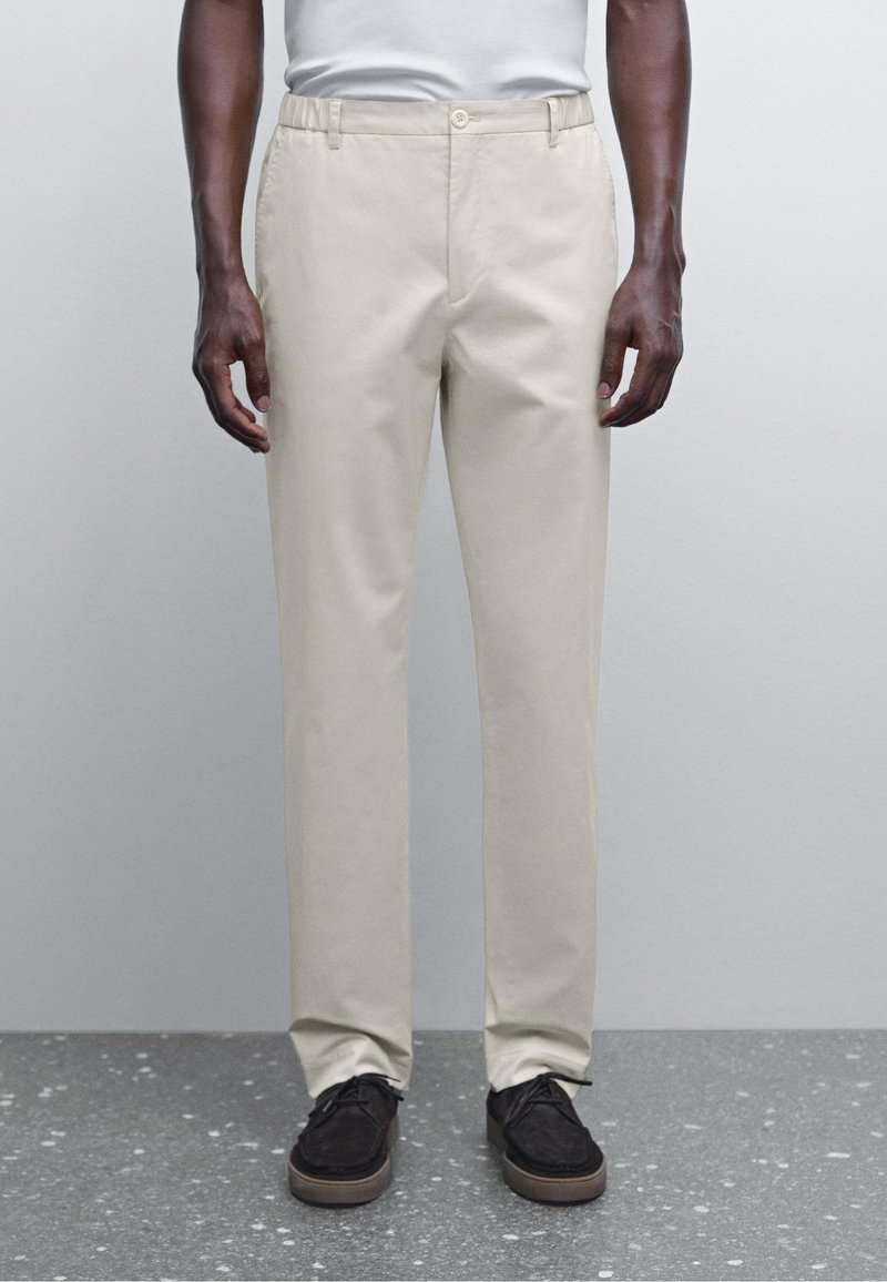 Homme portant un pantalon beige droit et des chaussures décontractées noires, debout contre un mur gris et un sol moucheté.