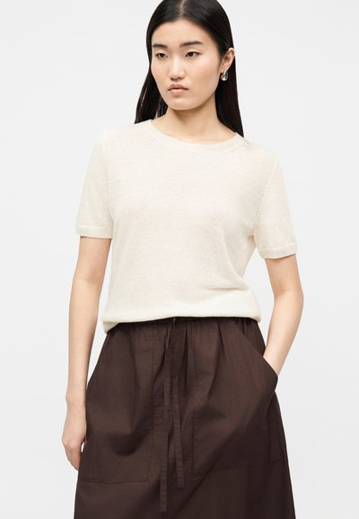 Jeune femme aux longs cheveux bruns portant un pull crème à manches courtes et une jupe taille haute marron foncé, les mains dans les poches.