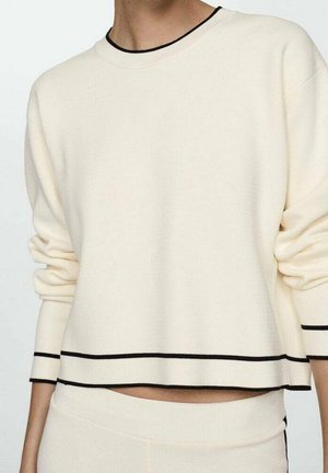Personne portant un pull crème à manches longues et un pantalon, tous deux avec de fines rayures noires aux poignets, à l'encolure et à l'ourlet.