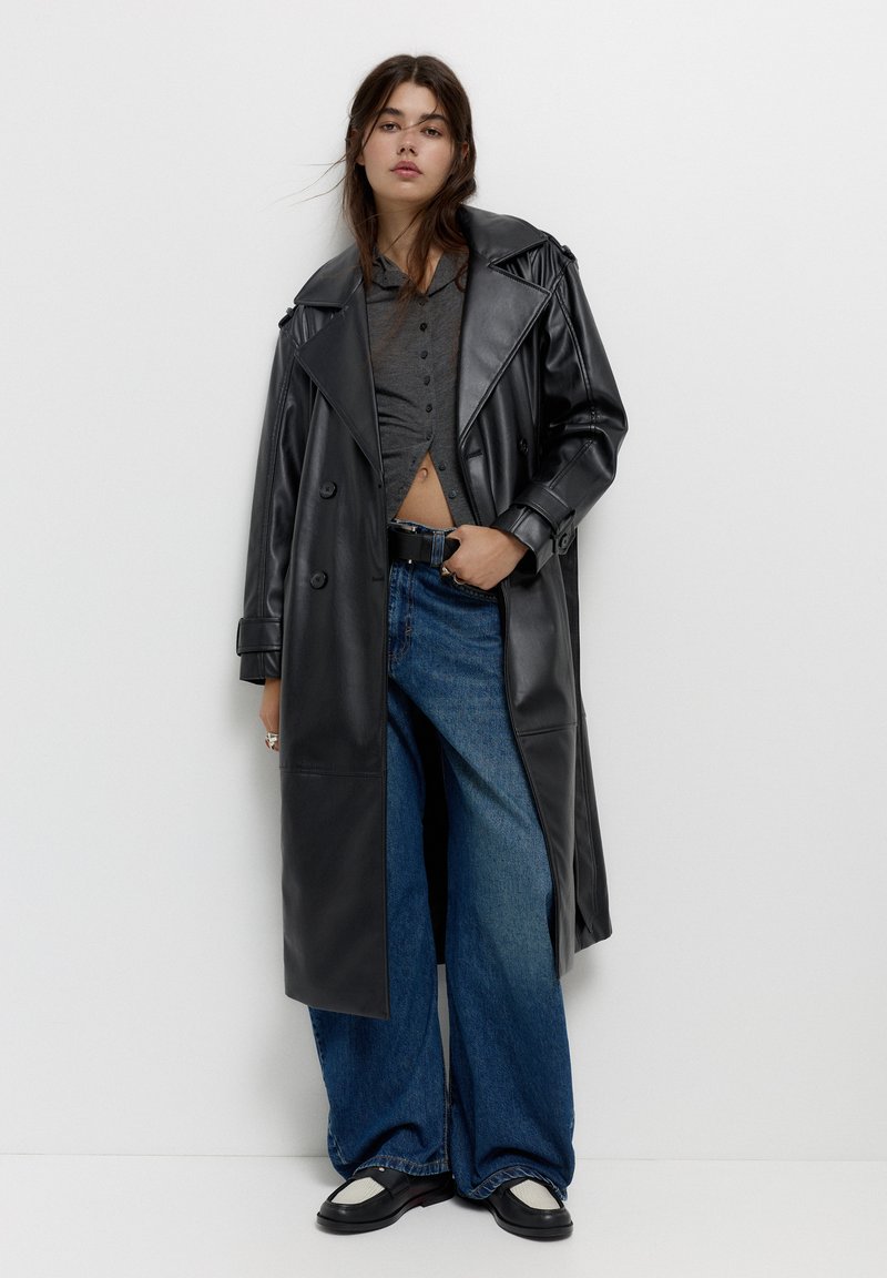 Femme portant un long manteau en cuir noir, une chemise courte à boutons gris foncé, un jean bleu large et des mocassins noir et blanc contre un mur blanc.