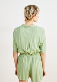 Rompère verte à manches courtes avec un tissu texturé, taille élastique et détail de bouton au dos. Collier lisse et coupe décontractée.