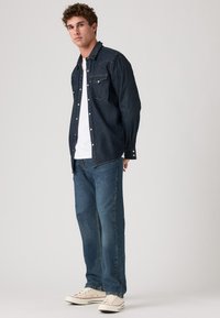 Donkerdenim overhemd met knopen en twee borstzakken, gecombineerd met lichtblauwe losvallende jeans en witte sneakers. Effen wit t-shirt eronder.