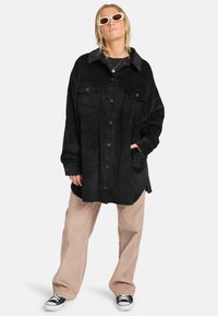 Schwarze Cordjacke mit Knopfleiste, Brusttaschen und einem kontrastierenden grauen Kragen. Kombiniert mit beigen Cordhosen und Sneakern.