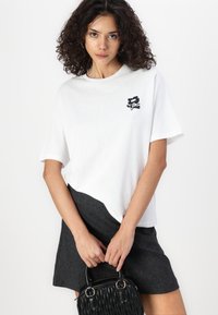 T-shirt en coton blanc avec un graphique noir sur la poitrine, porté avec une jupe gris foncé. Le modèle tient un petit sac à main noir texturé.