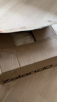 Kartonglåda delvis öppen, med en beige föremål inuti. Lådan har Zalando-logotypen i svart på sidan och är förseglad med transparent tejp.